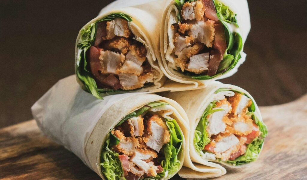 Four chicken shawarma wraps filled with crispy chicken, lettuce, and tomatoes are stacked neatly on a wooden surface.