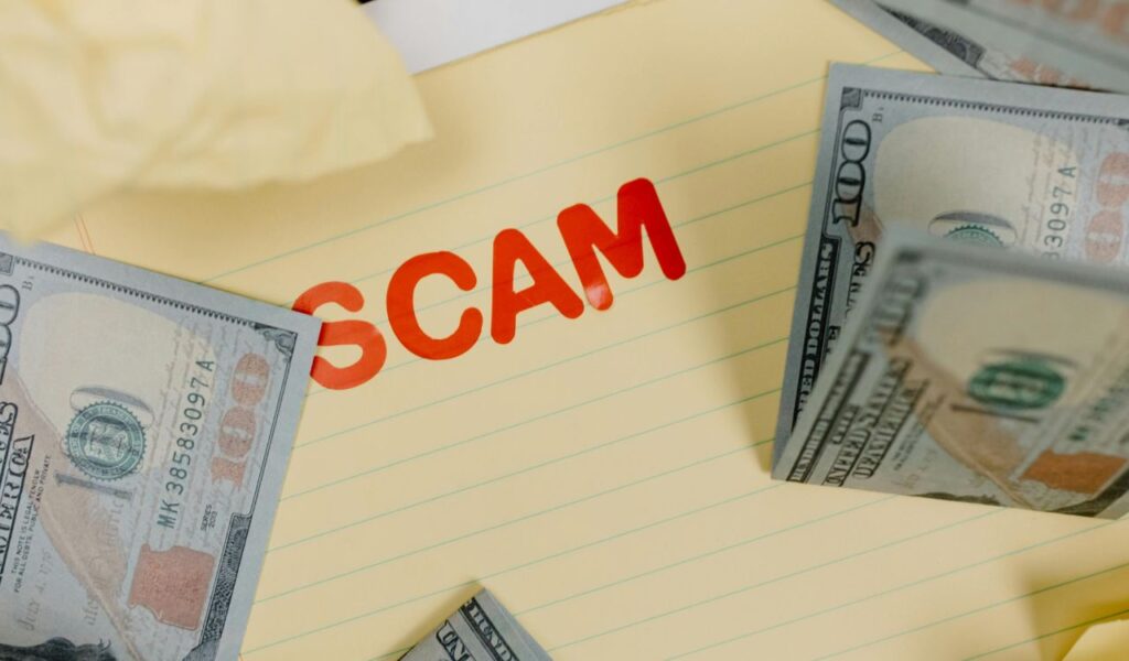 A stack of US 100-dollar bills surrounds a yellow notepad with the word "SCAM" written in bold red letters, highlighting the concept of a financial scam.