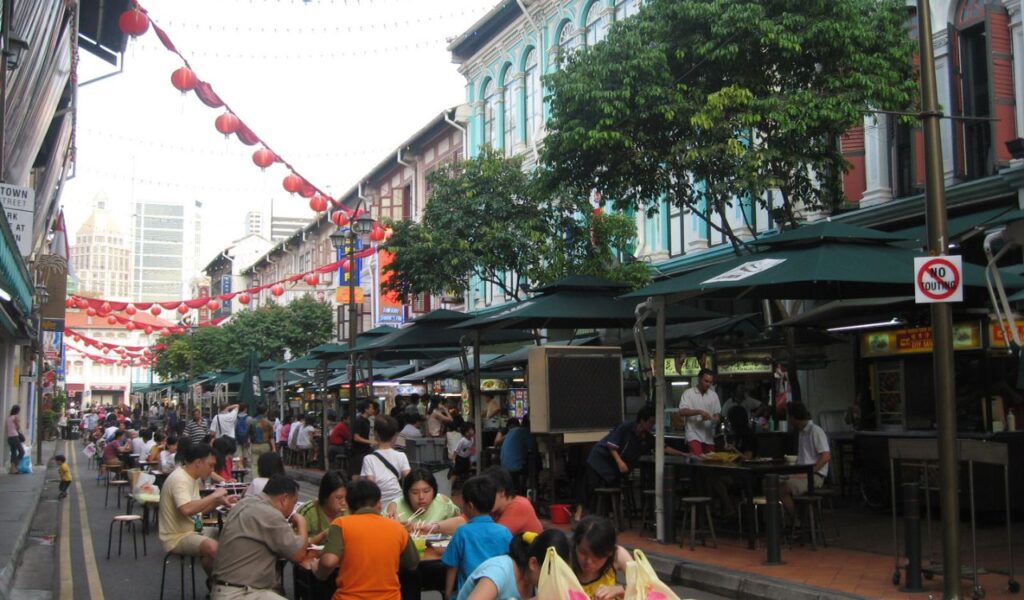 Busy Singapore Chinatown food street with outdoor diners, food stalls, red lanterns, and colorful colonial-style buildings.