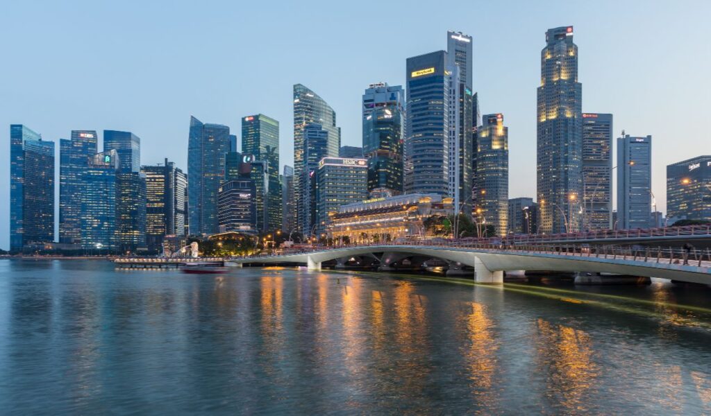 Singapore skyline at dusk with towering skyscrapers, illuminated lights, and a bridge crossing the tranquil river