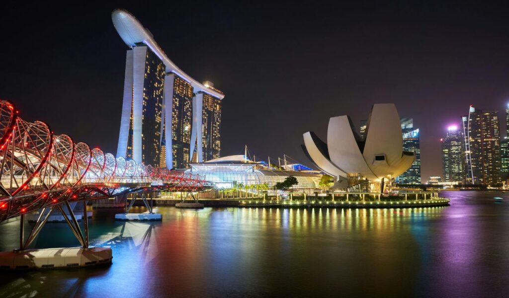 Night view of Marina Bay Sands and the ArtScience Museum in Singapore, with the Helix Bridge illuminated in red and blue lights over the water.