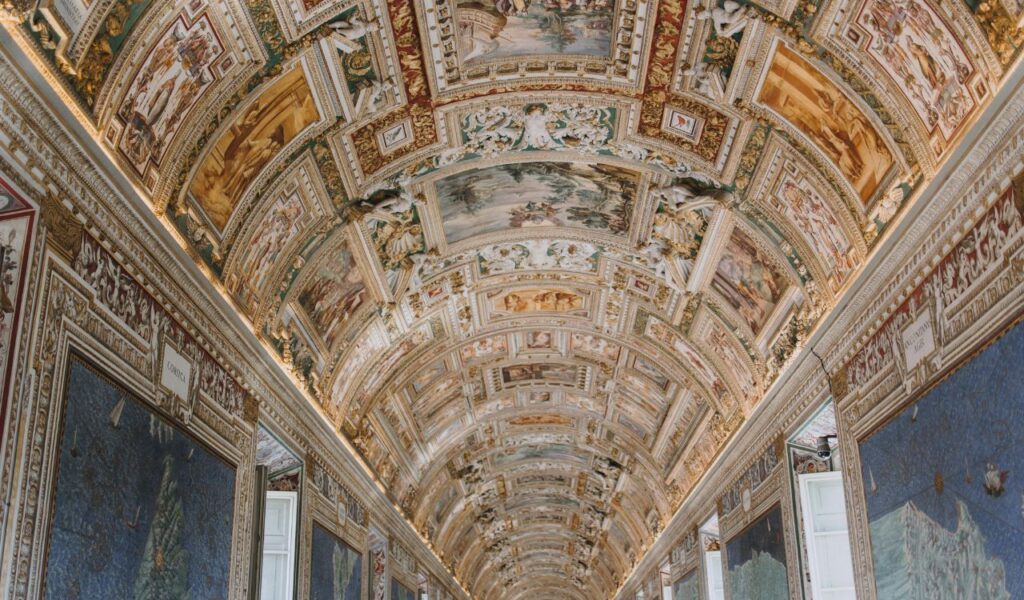 Ornate vaulted ceiling of the Sistine Chapel corridor adorned with intricate frescoes, gold detailing, and elaborate artwork depicting biblical scenes.
