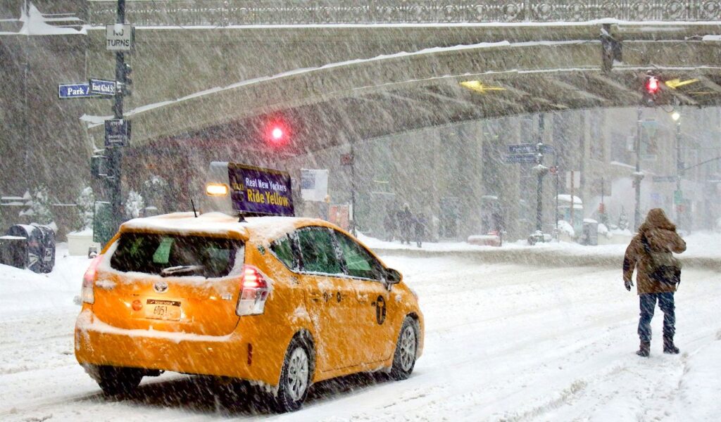 Yellow taxi driving cautiously through heavy snow in New York City while a pedestrian walks nearby in harsh weather.