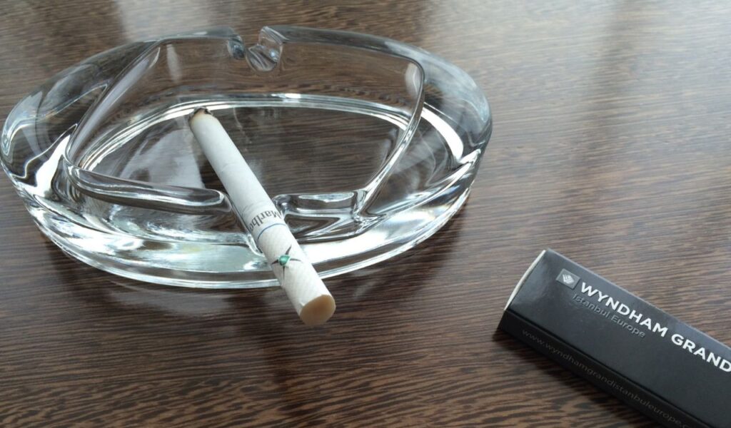 A cigarette rests in a clear glass ashtray on a wooden table, with a Wyndham Grand matchbox nearby