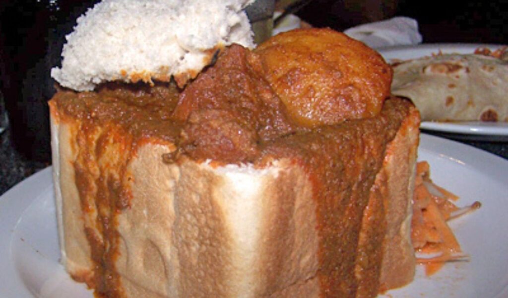 South African bunny chow with spicy curry filling inside a hollowed-out loaf of white bread, served on a white plate.