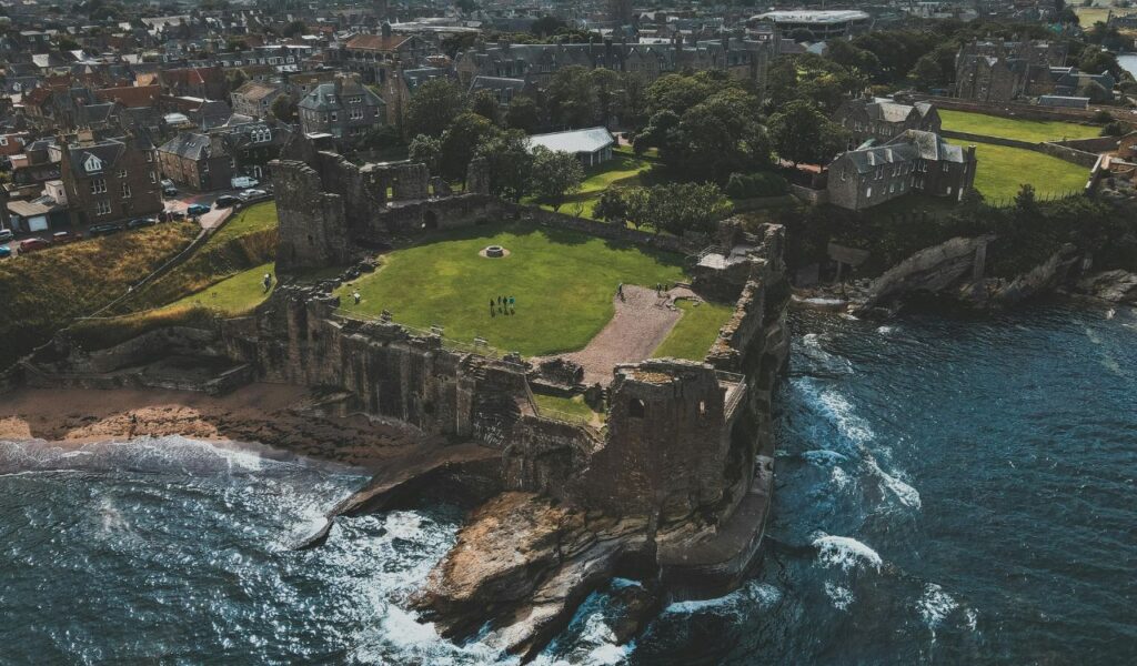 Aerial view of historic St. Andrews ruins perched on a rocky coastline, overlooking the ocean with a nearby town in the background.