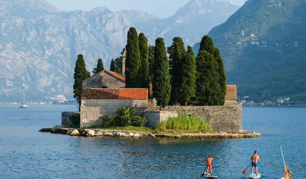 Stone building with red-tiled roofs and tall cypress trees on St. George Island, surrounded by blue waters and mountains, with paddleboarders in the foreground.