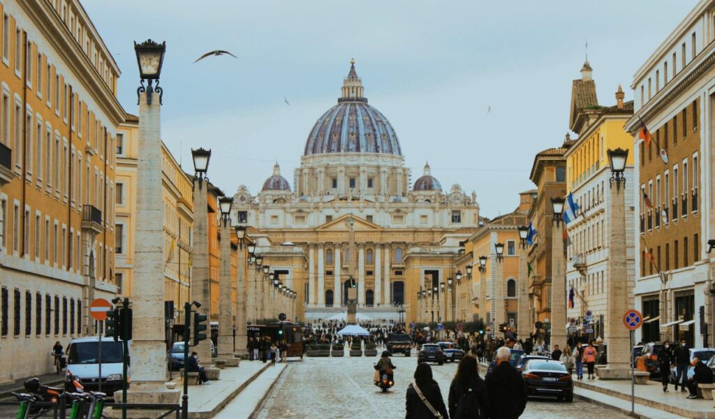View of St. Peter's Basilica from a cobblestone street lined with historic buildings, pedestrians, and vehicles in Vatican City.