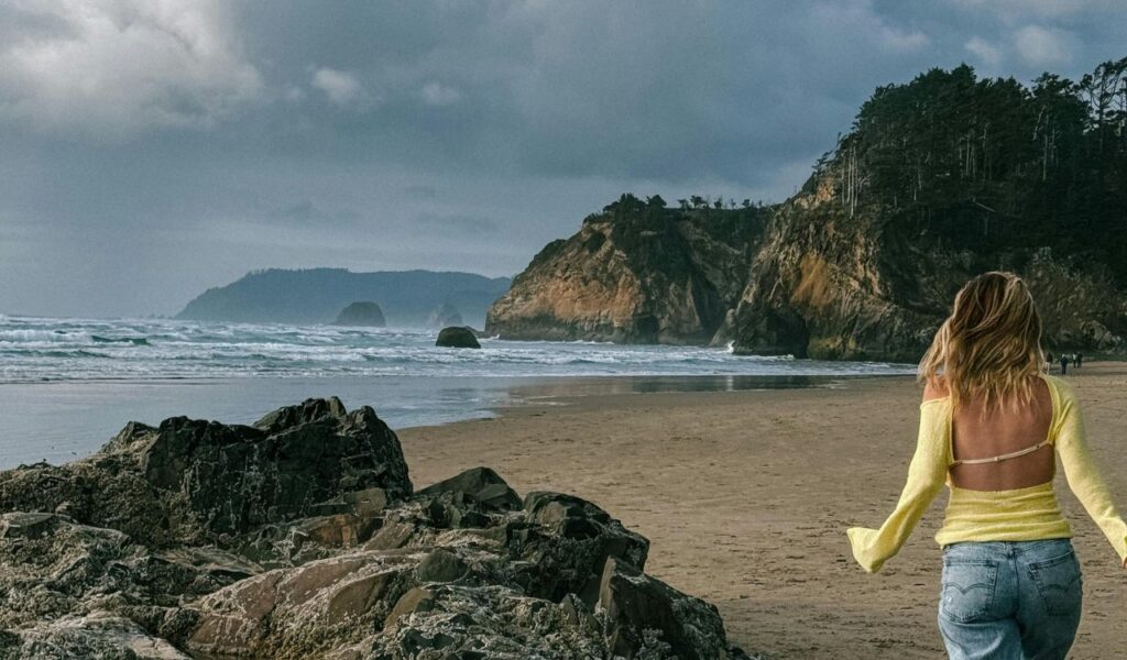 Woman walking alone on a rocky beach near cliffs and ocean waves, symbolizing slow travel and extended stays.