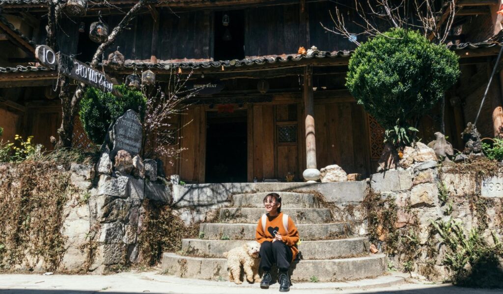 Person sitting on stone steps with a small dog in front of a rustic wooden homestay surrounded by plants and decorative signs.