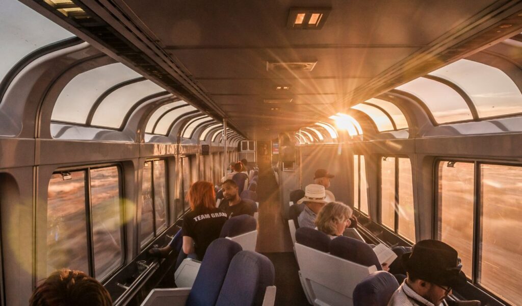 Passengers relaxing in a train observation car with large panoramic windows, enjoying sunset views over the landscape outside.