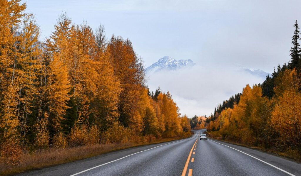 A car driving along a scenic highway surrounded by autumn-colored trees, with snow-capped mountains and low clouds in the distance.
