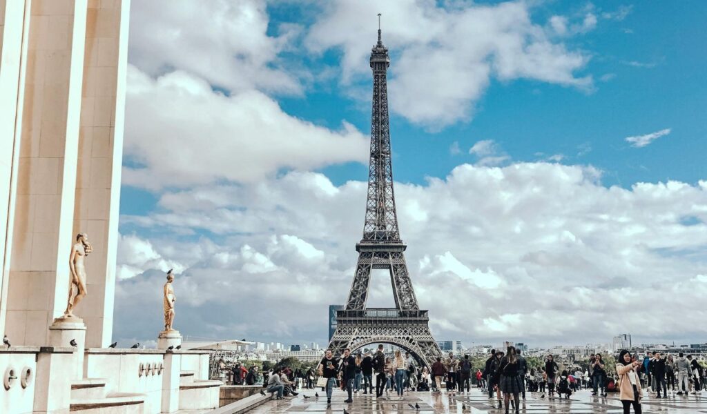 Tourists gather near the Eiffel Tower on a bright day with scattered clouds, enjoying the iconic Paris landmark and lively surroundings.