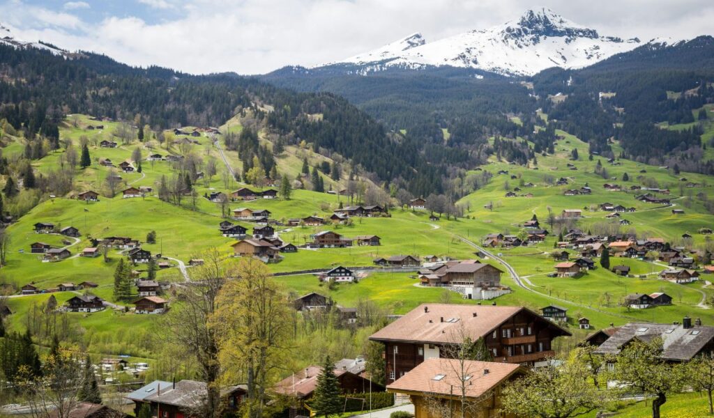 A scenic Swiss village with wooden houses, green hills, and snow-capped mountains.