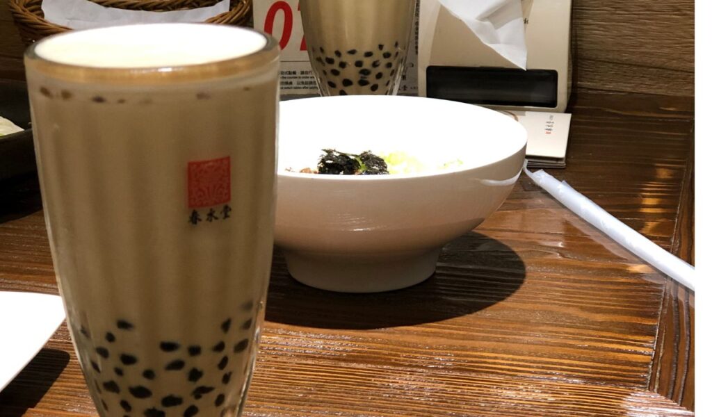 Glass of Taiwanese bubble tea with black tapioca pearls on a wooden table, served next to a white bowl of food and napkins.