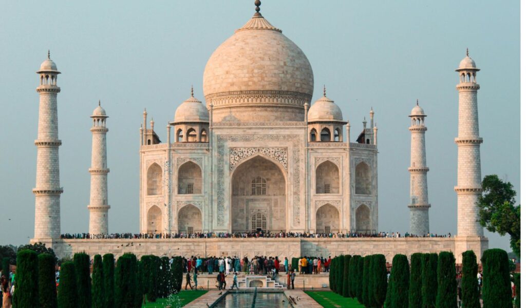 The Taj Mahal in India with its white marble dome and minarets, surrounded by gardens and reflecting pools with crowds of visitors.