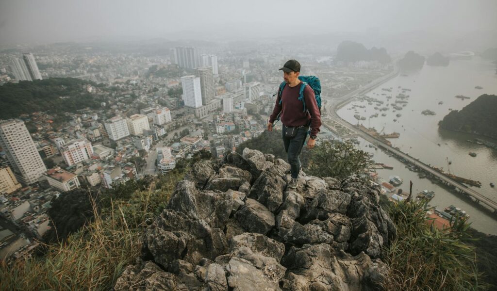 A solo backpacker hikes a rocky ridge above a sprawling city and harbor, taking in the slow beauty of overland travel.