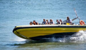 Group of people wearing life vests riding a yellow jet boat on a river, with water splashing around the boat and a guide steering at the back.