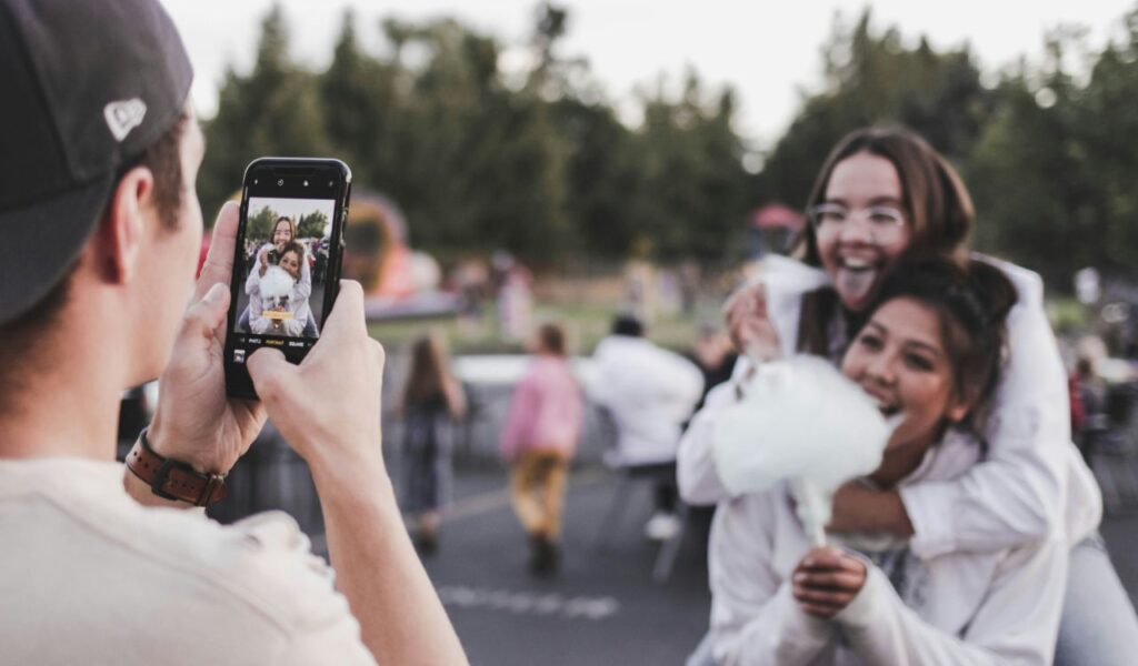 Person taking a photo with a smartphone of two smiling friends, one holding cotton candy, at an outdoor event with a blurred crowd in the background