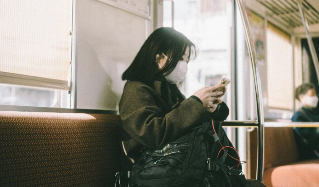 A young woman wearing a face mask sits quietly on a train in Japan, looking at her phone with a black backpack in her lap.