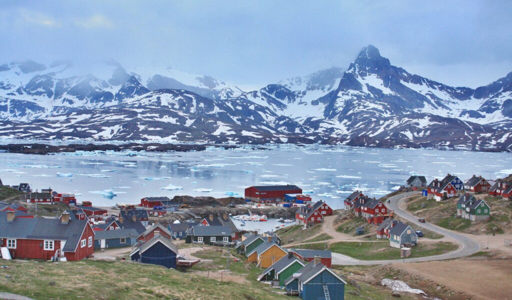A coastal Greenlandic village with colorful houses, icy waters, and snow-covered mountains in the background.