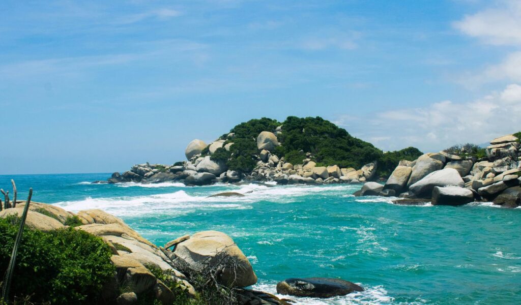 Tayrona National Park in Colombia with turquoise waters, rocky shoreline, and lush green vegetation under a bright sky.