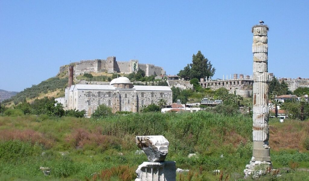 Ruins of the Temple of Artemis in Ephesus, Turkey, with a single standing column surrounded by greenery and historic buildings in the background, including a mosque and a medieval fortress