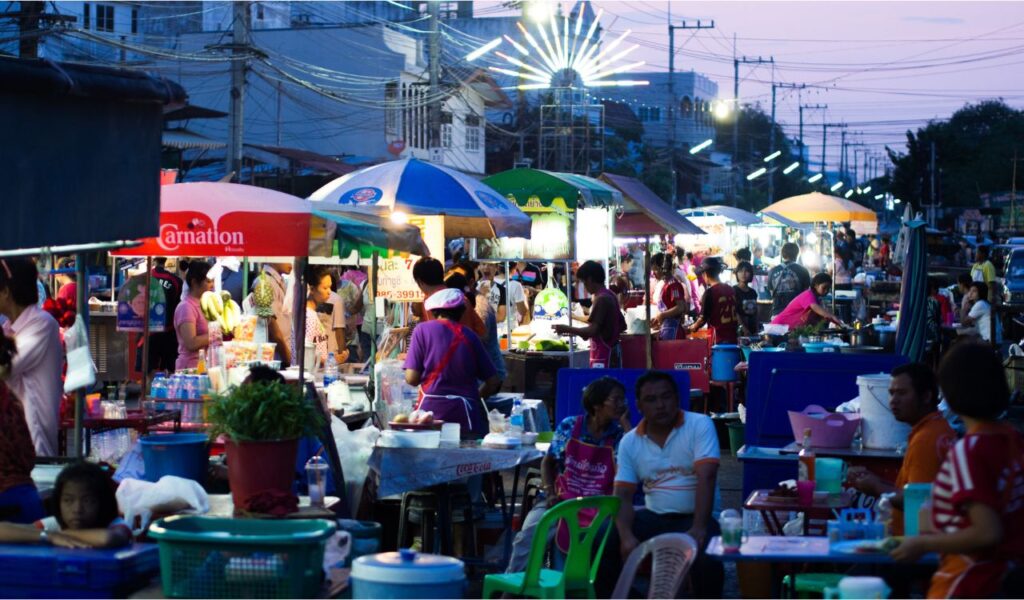 Busy night street food market in Thailand with locals and tourists enjoying traditional Thai dishes under colorful umbrella