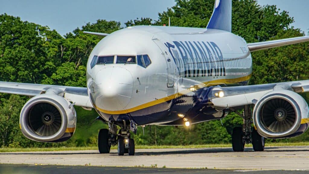 Close-up front view of Ryanair aircraft taxiing on runway with engines running, surrounded by lush green trees.