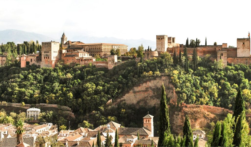 The Alhambra in Spain with its historic palaces and towers set atop a hill, surrounded by lush greenery and overlooking nearby houses.