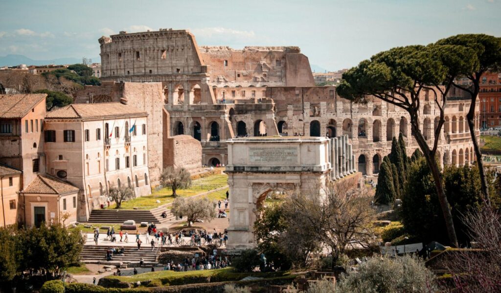 The Colosseum in Rome, Italy, with its iconic ancient stone arches surrounded by nearby buildings, trees, and crowds of visitors.