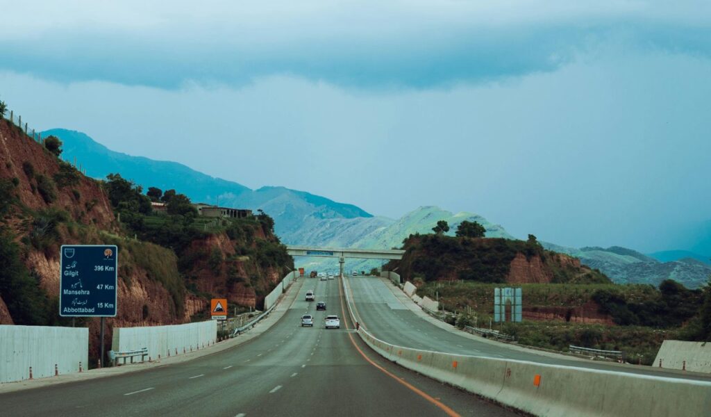 Wide highway cutting through green hills and red cliffs on The Great Northern (US-2), with distant mountains under a cloudy blue sky.