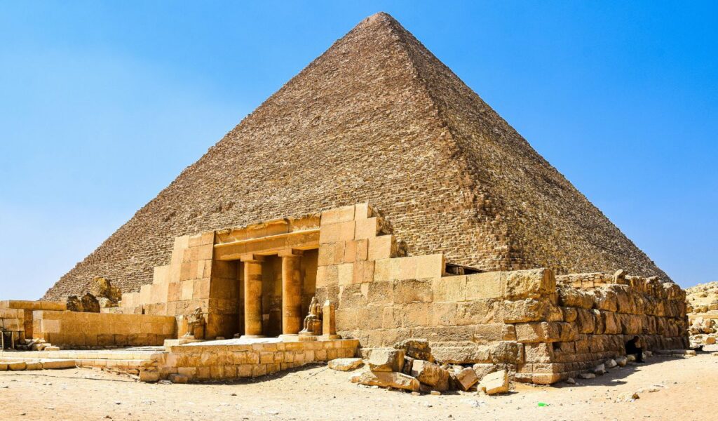 The Great Pyramid of Giza in Egypt under a clear blue sky, showing its massive triangular structure made of stone blocks with an ancient entrance and surrounding ruins in the foreground.