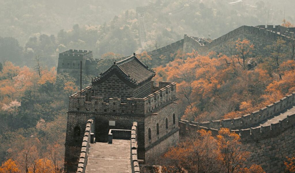 The Great Wall of China winding over hills with a watchtower in view, surrounded by autumn trees with orange foliage.