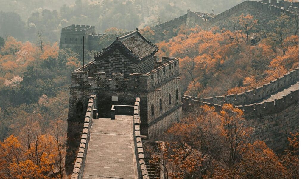 The Great Wall of China winding over hills with a watchtower in view, surrounded by autumn trees with orange foliage.