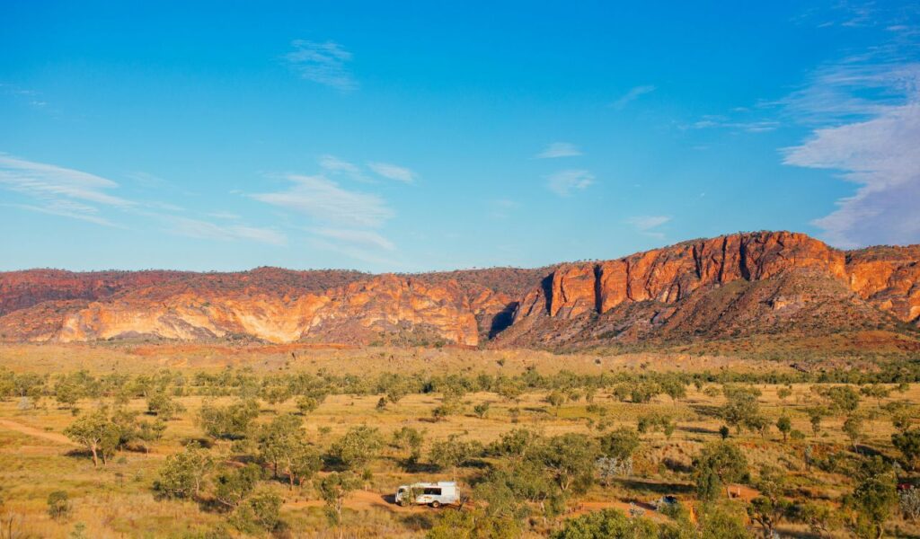 A lone camper van drives through Australia’s rugged Outback near orange cliffs under a wide open blue sky
