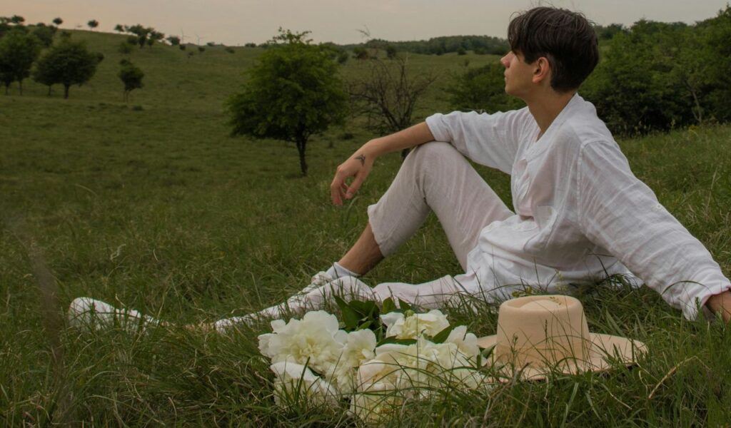 A person dressed in white relaxes on a grassy hill surrounded by flowers, enjoying a peaceful moment in nature with scenic views.