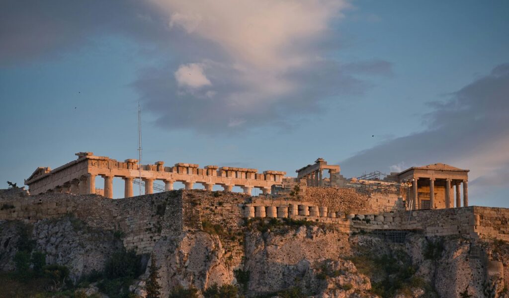 The Parthenon in Greece atop a rocky hill, featuring ancient columns and partially ruined structures under a cloudy sky at sunset.