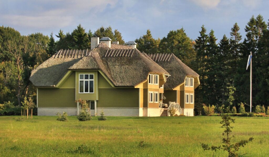A large countryside guesthouse with a thatched roof sits quietly in a sunny open field, surrounded by trees and greenery.