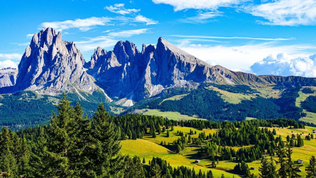 **Alt text:**
Panoramic view of the Dolomites with jagged peaks rising above a lush green valley dotted with pine trees and scattered cabins under a bright blue sky with clouds.