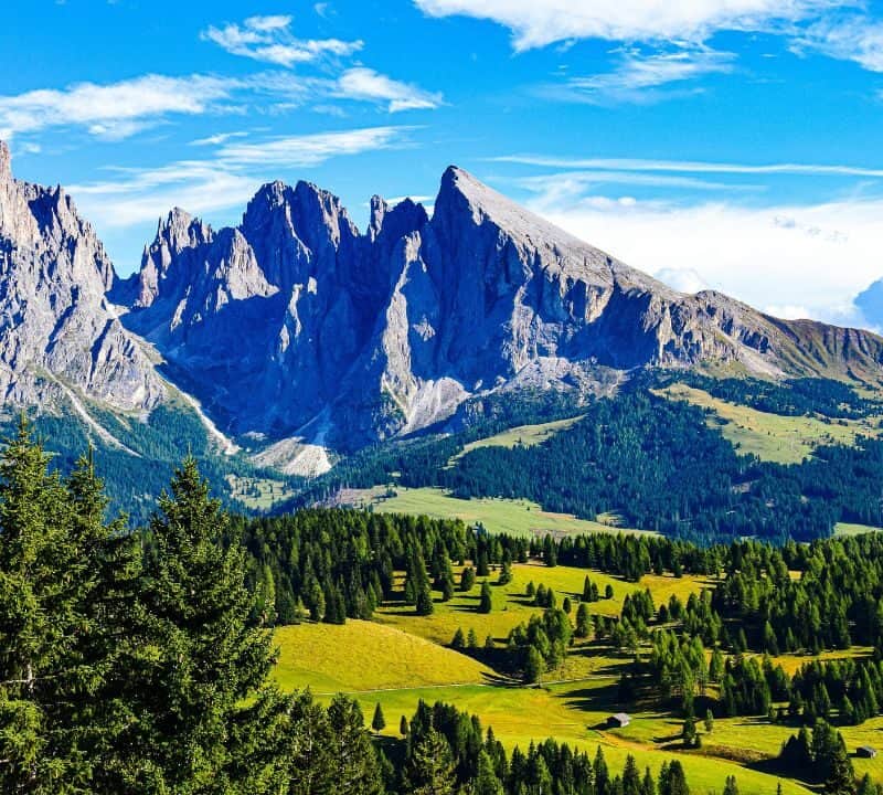 **Alt text:** Panoramic view of the Dolomites with jagged peaks rising above a lush green valley dotted with pine trees and scattered cabins under a bright blue sky with clouds.