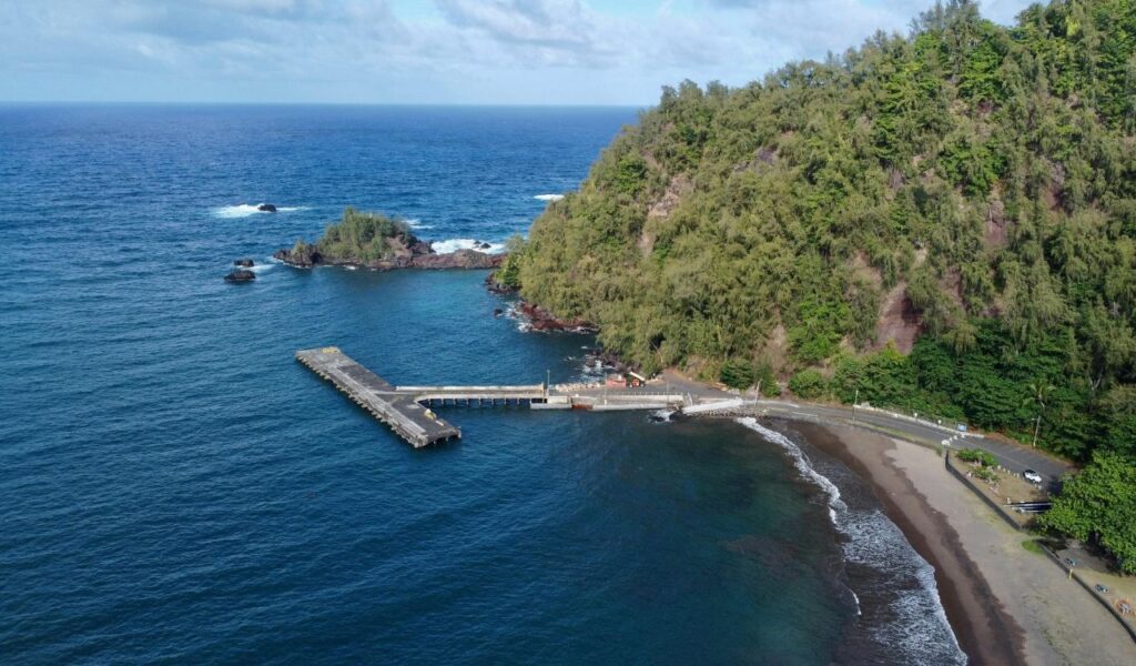 Aerial view of the Road to Hana in Hawaii, with a coastal pier, lush green cliffs, and deep blue ocean waters under a partly cloudy sky.