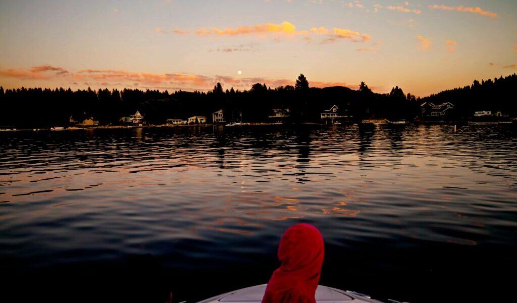 Person in a red hoodie sits at the front of a boat, looking across a calm lake toward shoreline houses during a colorful sunset.