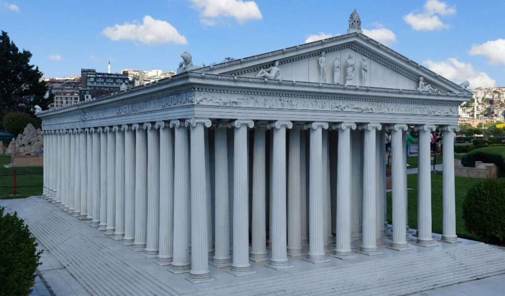 he Temple of Artemis at Ephesus with tall Ionic columns and carved details, set in a grassy area under a blue sky.