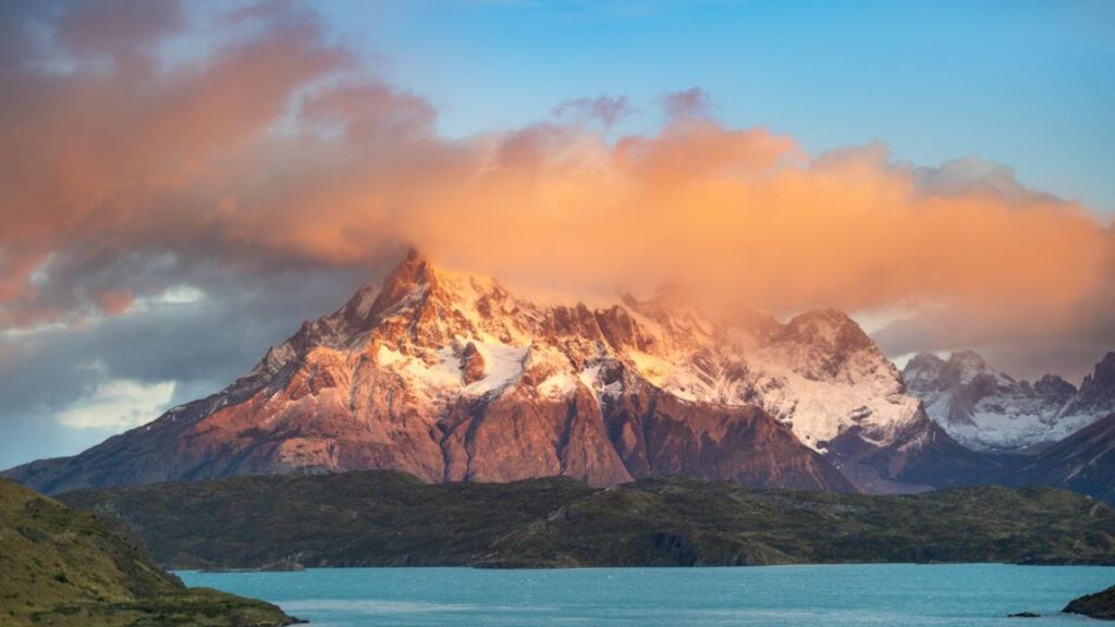 Snow-capped mountain peaks in South America at sunset, with vibrant orange clouds above and a turquoise lake in the foreground.