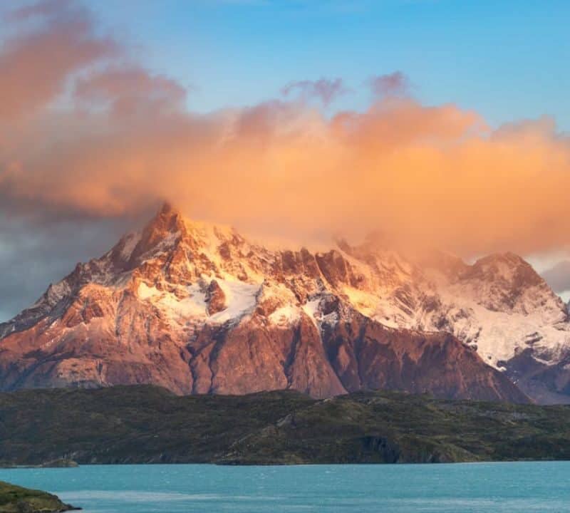 Snow-capped mountain peaks in South America at sunset, with vibrant orange clouds above and a turquoise lake in the foreground.