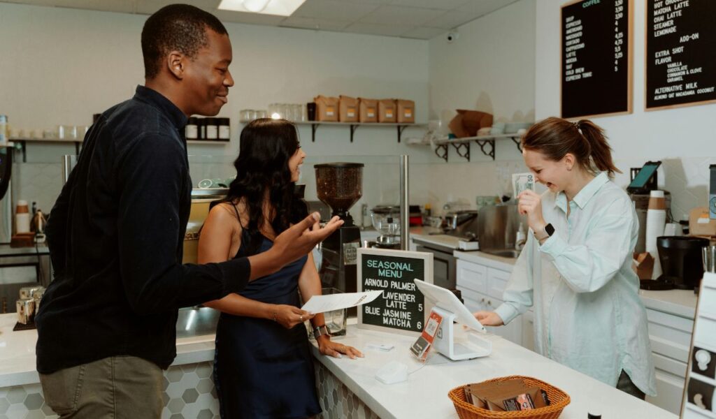 o customers smiling and talking with a cashier while making a payment at a coffee shop counter.