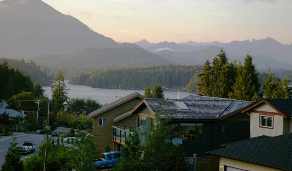 Scenic view of Tofino, Canada, with forested mountains, calm waters, sailboats, and coastal homes at sunset.