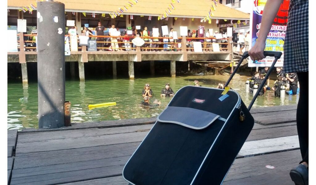 Tourist pulling luggage on a pier near a crowd and scuba divers, possibly at a tourist hotspot or ferry terminal.