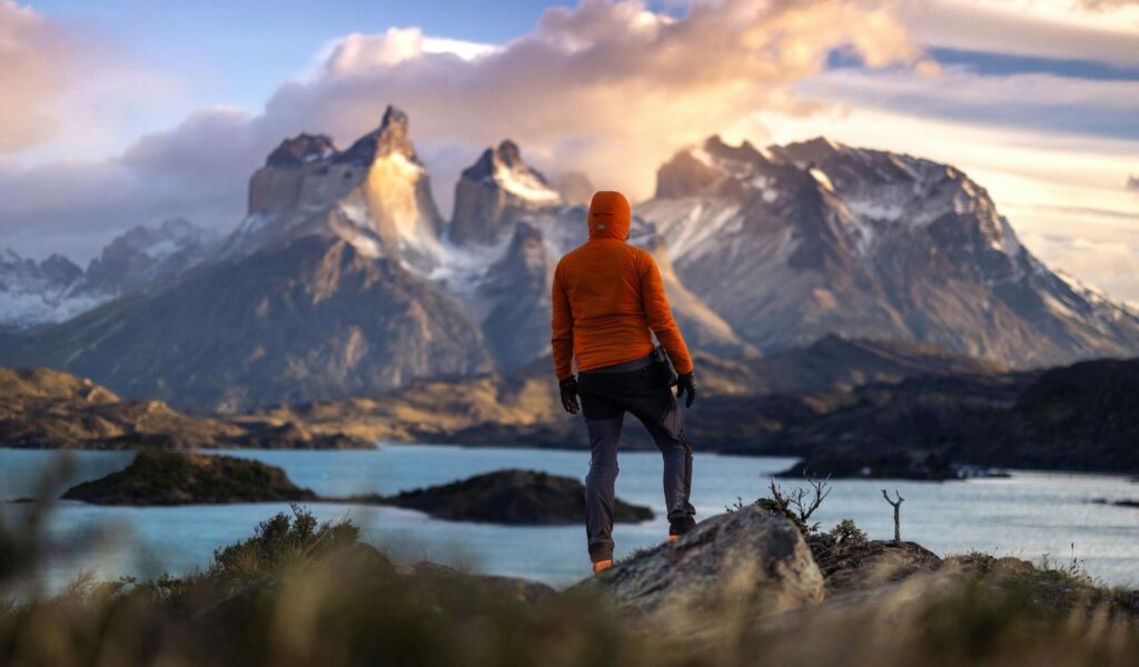 A hiker in an orange jacket stands overlooking Torres del Paine in Chile, with rugged mountains and blue lakes under a dramatic sky.